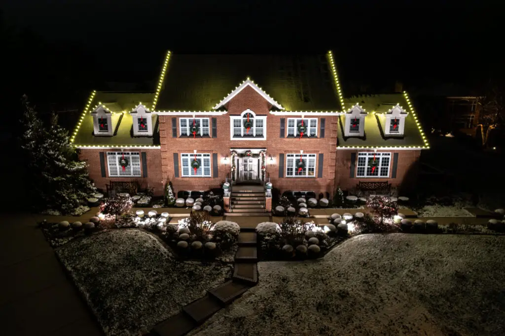 Drone view of a stately brick home featuring elegant warm white holiday lighting professionally installed by Washington Holiday Lighting. Custom LED roofline lights, illuminated landscaping, festive wreaths, and seasonal garland create a timeless residential Christmas display that enhances curb appeal and showcases expert outdoor holiday lighting installation in a snow-covered winter setting.