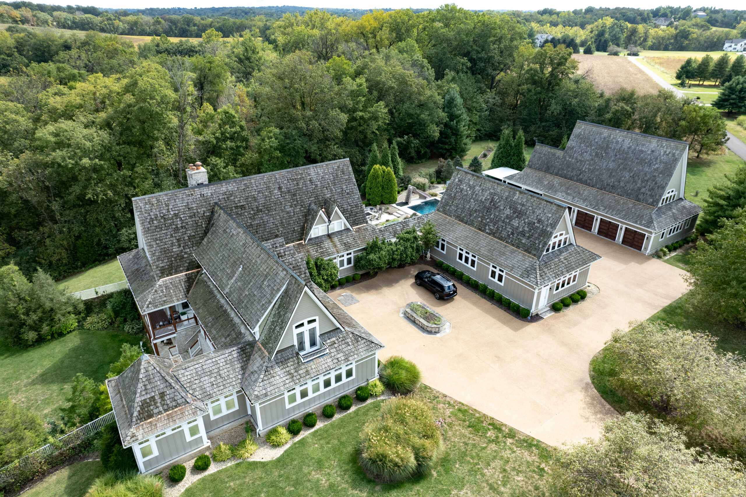 **Alt Text:** Aerial drone photograph of a large custom residential estate surrounded by mature trees and open countryside. The property features multiple interconnected buildings with steeply pitched roofs clad in weathered cedar shake shingles, creating a cohesive architectural design. A spacious concrete driveway leads to an attached garage and courtyard area, where a single vehicle is parked. In the center of the property, a backyard pool and outdoor living space are visible between the main house and secondary structures. The home is set on a well-maintained landscaped lot with manicured shrubs, expansive lawns, and wooded surroundings, offering privacy and scenic rural views.