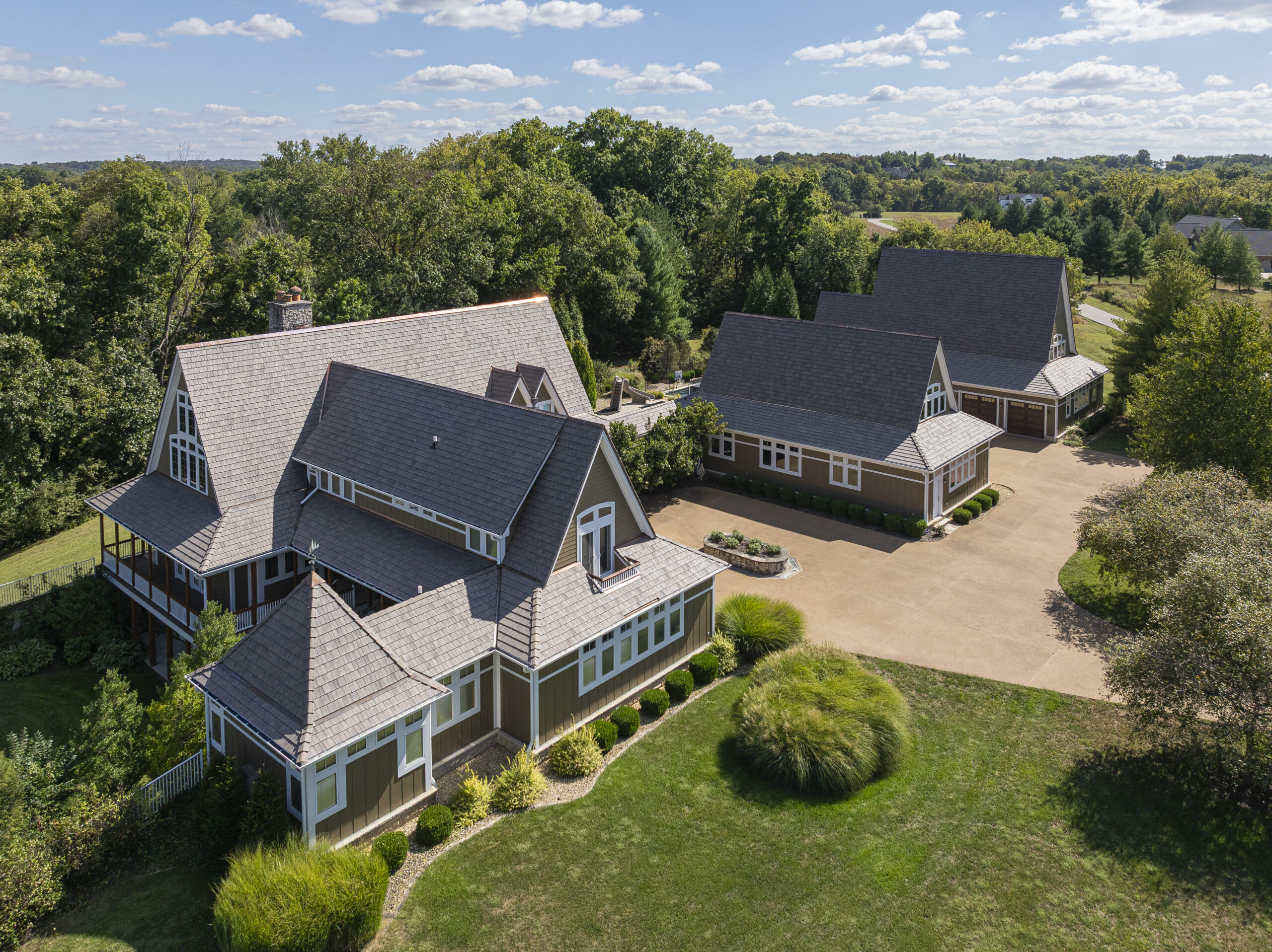 **Alt Text:** Aerial drone photograph of a large luxury residential estate featuring a newly installed composite shake-style roofing system from davinci. The property includes multiple interconnected structures with steeply pitched rooflines, dormers, and decorative architectural details, all finished with a uniform weathered-gray roof. A spacious concrete driveway connects the main residence to a detached garage and auxiliary building, creating a courtyard-style layout. The home is surrounded by expansive lawns, landscaped planting beds, mature trees, and rolling countryside views. The image showcases the scale, craftsmanship, and curb appeal of the completed roofing project while highlighting the property's rural setting and well-maintained grounds.