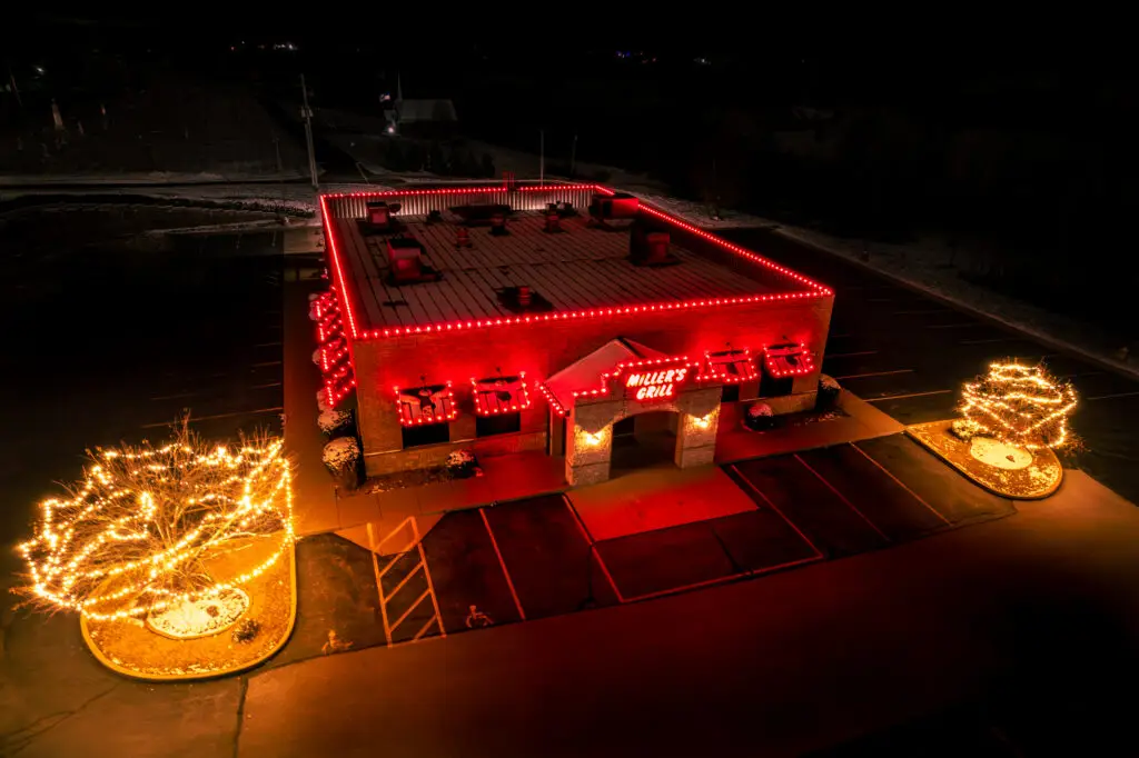 Drone view of a commercial restaurant featuring professional holiday lighting with vibrant red LED roofline lights and warmly illuminated trees. This custom commercial holiday lighting installation enhances the building's exterior, creating a festive and inviting Christmas display that attracts customers and showcases professional outdoor holiday lighting design.