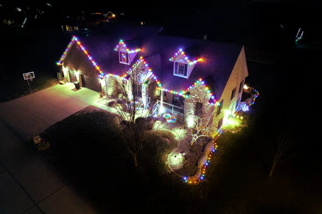 - Aerial nighttime view of a large custom home featuring professionally installed multicolor Custom cut Christmas lights outlining the roofline, dormers, front porch, and landscape borders, creating a vibrant and elegant residential holiday lighting display.