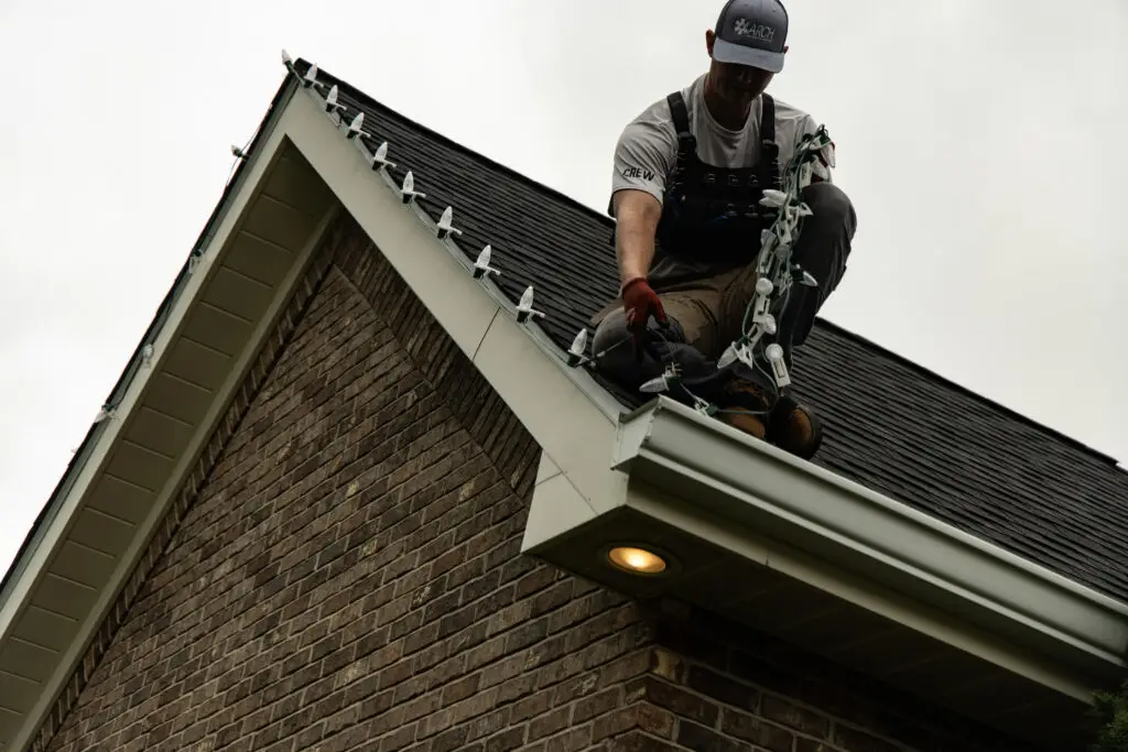 - Holiday lighting installer kneeling on the edge of a residential roof while securing white C9 Christmas lights along the gutterline of a brick home, demonstrating professional roofline holiday light installation.