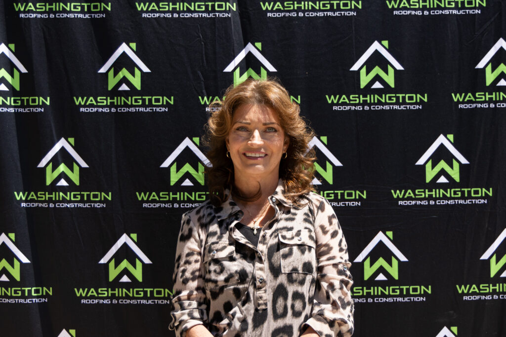 Washington Roofing & Construction team member Tacia Biene- sales assistant. standing in front of a backdrop featuring the logo and name "Washington Roofing & Construction." She is wearing a patterned blouse and smiling at the camera.