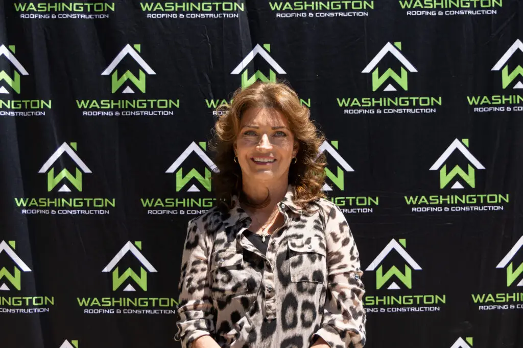 Washington Roofing & Construction team member Tacia Biene- sales assistant. standing in front of a backdrop featuring the logo and name "Washington Roofing & Construction." She is wearing a patterned blouse and smiling at the camera.